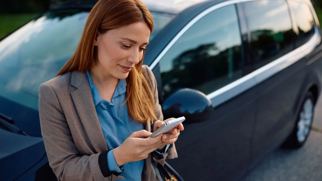 Eine rothaarige Frau lehnt an ihrem Auto oder Mietwagen und schaut auf ihr Handy.