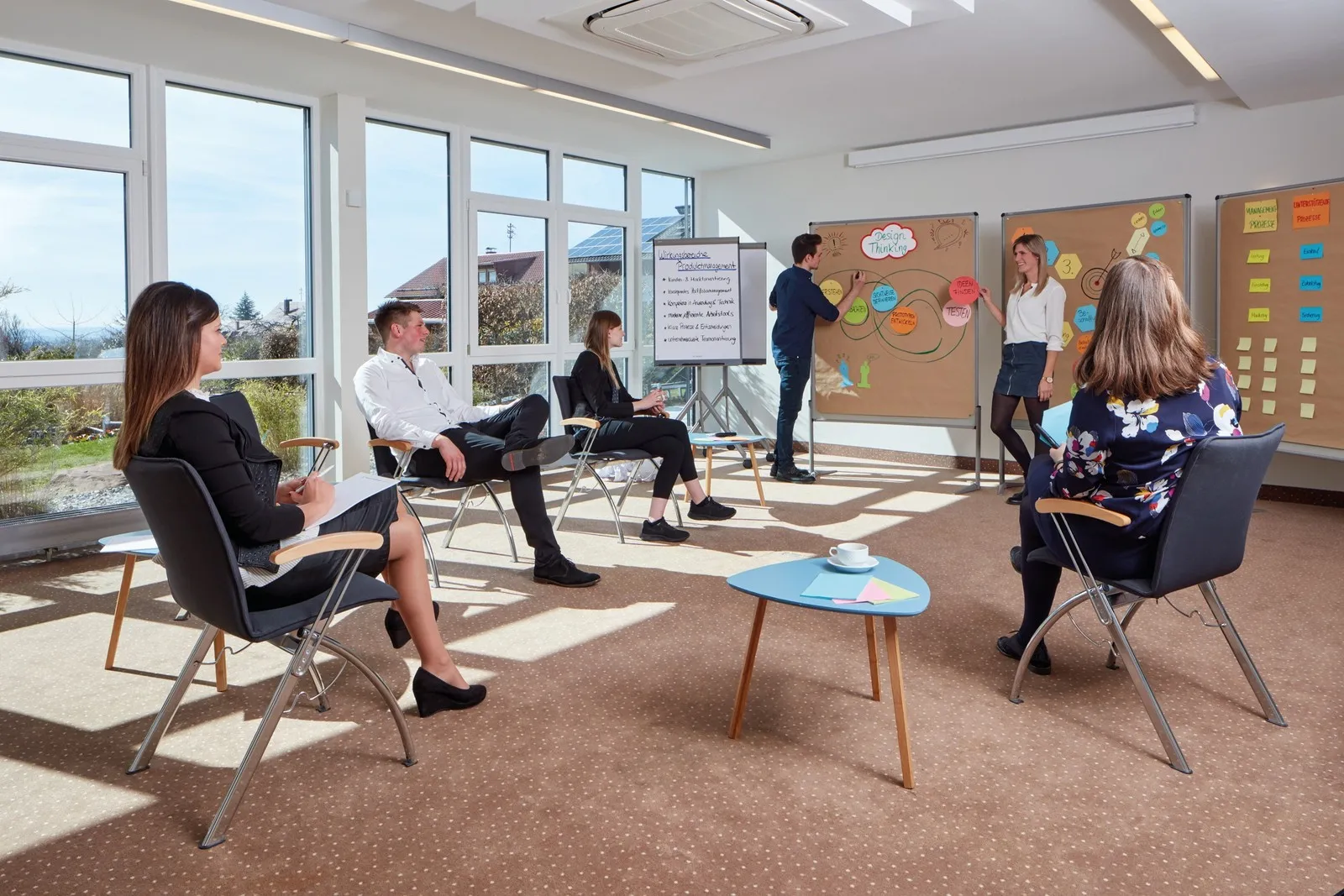 Eine Gruppe nimmt in einem hellen Seminarraum an einem Workshop teil. Zwei Personen stehen an Pinnwänden mit Design-Thinking-Notizen, weitere Teilnehmende sitzen im Stuhlkreis.