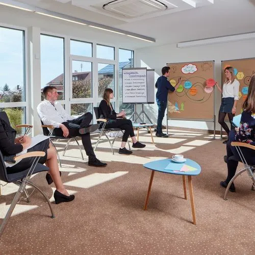Eine Gruppe nimmt in einem hellen Seminarraum an einem Workshop teil. Zwei Personen stehen an Pinnwänden mit Design-Thinking-Notizen, weitere Teilnehmende sitzen im Stuhlkreis.