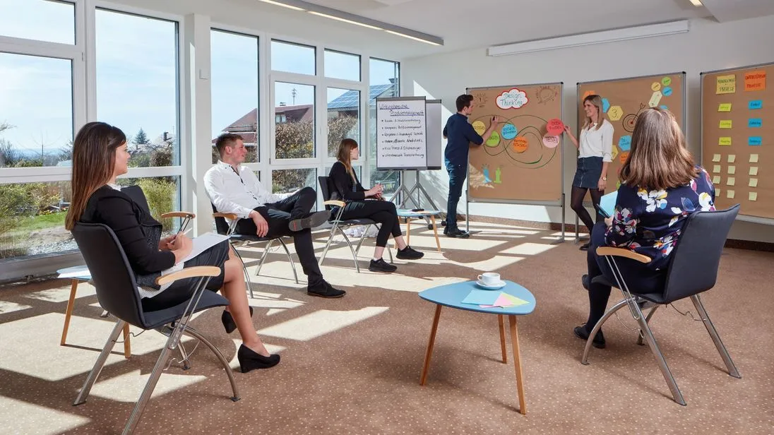 Eine Gruppe nimmt in einem hellen Seminarraum an einem Workshop teil. Zwei Personen stehen an Pinnwänden mit Design-Thinking-Notizen, weitere Teilnehmende sitzen im Stuhlkreis.