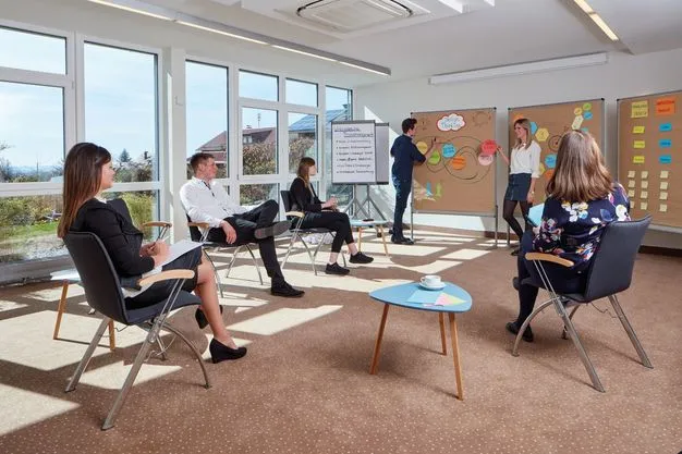 Eine Gruppe nimmt in einem hellen Seminarraum an einem Workshop teil. Zwei Personen stehen an Pinnwänden mit Design-Thinking-Notizen, weitere Teilnehmende sitzen im Stuhlkreis.