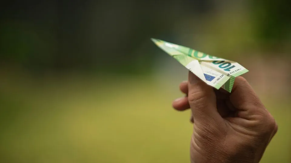 Eine Hand hält ein aus einem 100-Euro-Schein gefaltetes Papierflugzeug vor unscharfem Hintergrund.