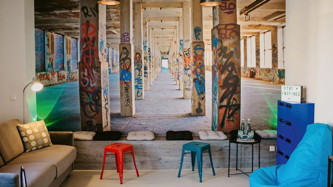 Ein Aufenthaltsraum mit Sitzgelegenheiten, farbigen Hockern und einer großen Wandaufnahme einer Industriehalle mit Graffiti.