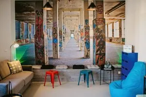Ein Aufenthaltsraum mit Sitzgelegenheiten, farbigen Hockern und einer großen Wandaufnahme einer Industriehalle mit Graffiti.