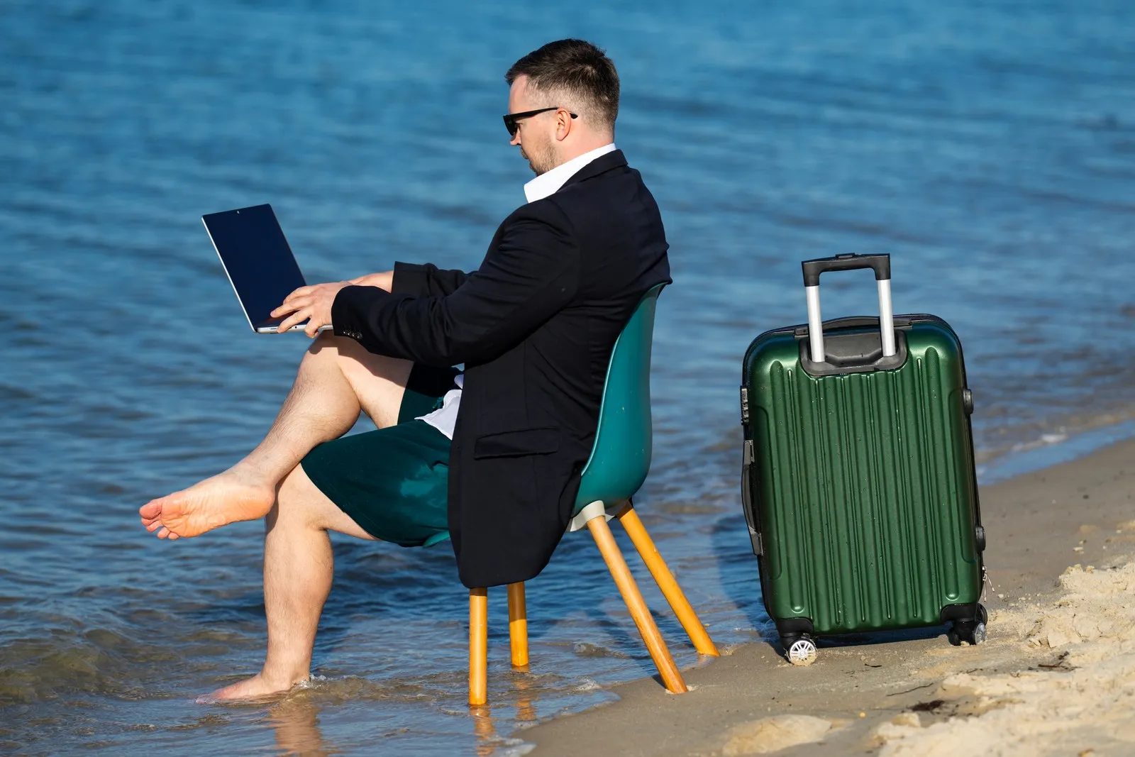 Ein Mann sitzt mit Koffer und Laptop auf dem Schoß am Strand und hat seine Füße im Meer.