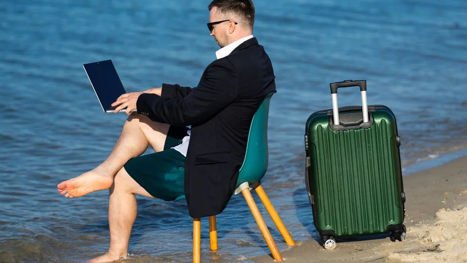 Ein Mann sitzt mit Koffer und Laptop auf dem Schoß am Strand und hat seine Füße im Meer.